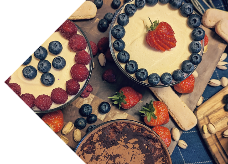 Top view of desserts arranged on a wooden surface, including tiramisu with blueberries and strawberries, and various cookies and nuts scattered around.